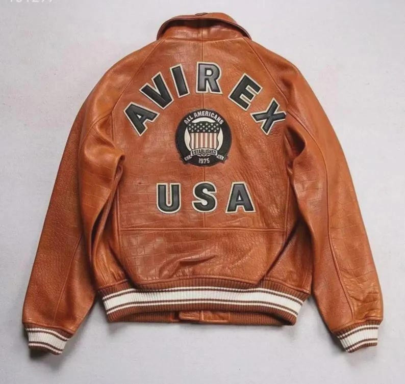 Pu&ograve; includere: Giacca in pelle marrone con la scritta "AVIREX" ad arco sulle spalle e "USA" sulla parte inferiore della schiena. Una toppa circolare con le parole "All Americans" e "Established 1975" &egrave; centrata sopra il testo "USA". La giacca ha polsini a righe bianche e nere.