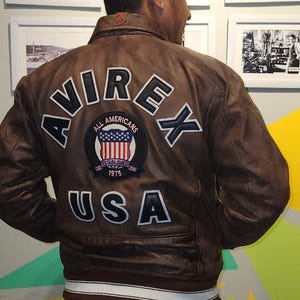 May include: Brown leather jacket with the word "AVIREX" across the back in white lettering. Below the word "AVIREX" is a patch with the words "ALL AMERICANS" and "ESTABLISHED 1975" with a USA flag design. The word "USA" is below the patch.