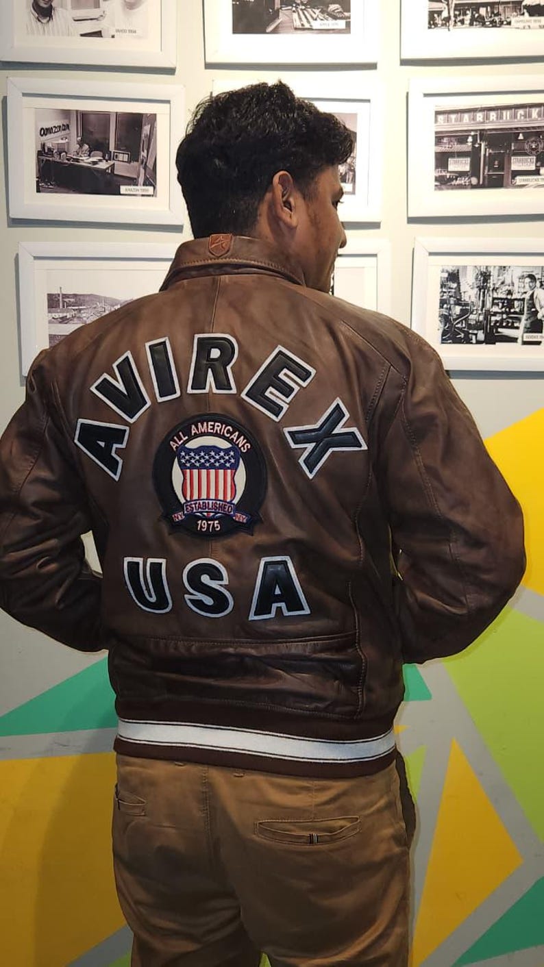 May include: Brown leather jacket with the word "AVIREX" across the back in white lettering. Below the word "AVIREX" is a patch with the words "ALL AMERICANS" and "ESTABLISHED 1975" with a USA flag design. The word "USA" is below the patch.