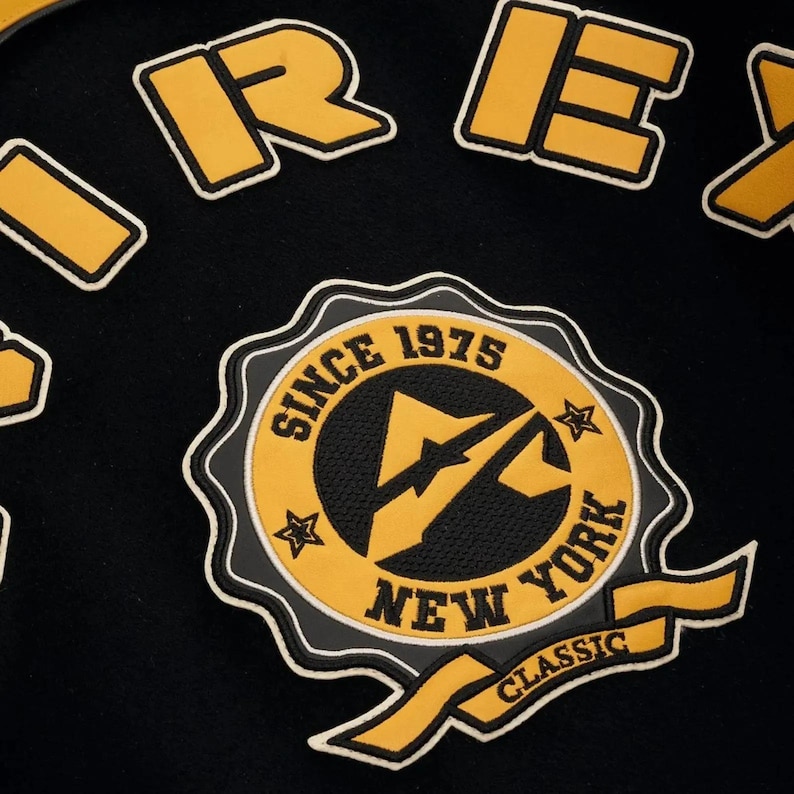 May include: Close-up of a black fabric with yellow and white embroidered patches. The patches include the words "REX" and a circular emblem with "SINCE 1975 NEW YORK CLASSIC" and a lightning bolt design.