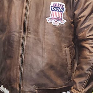 May include: Brown leather bomber jacket with a zip-up front, a collar, and a patch on the chest that reads "AVIREX ESTABLISHED 1975". The jacket has white stripes on the cuffs and hem.