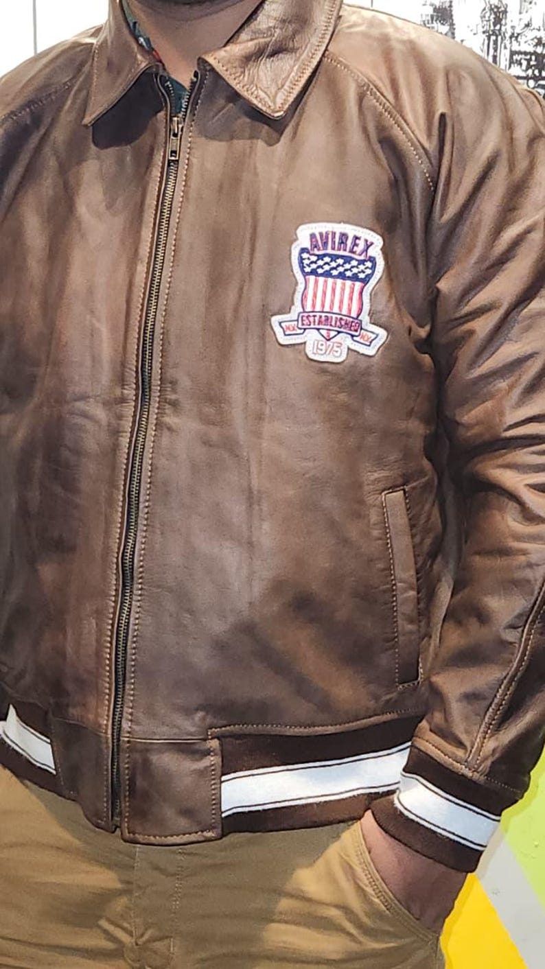 May include: Brown leather bomber jacket with a zip-up front, a collar, and a patch on the chest that reads "AVIREX ESTABLISHED 1975". The jacket has white stripes on the cuffs and hem.