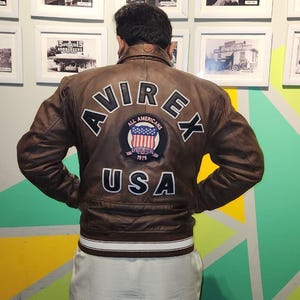 May include: A brown leather jacket with the word "AVIREX" in white lettering across the back, along with a patch that says "All Americans" and "USA". The jacket has a white and brown striped band at the waist.