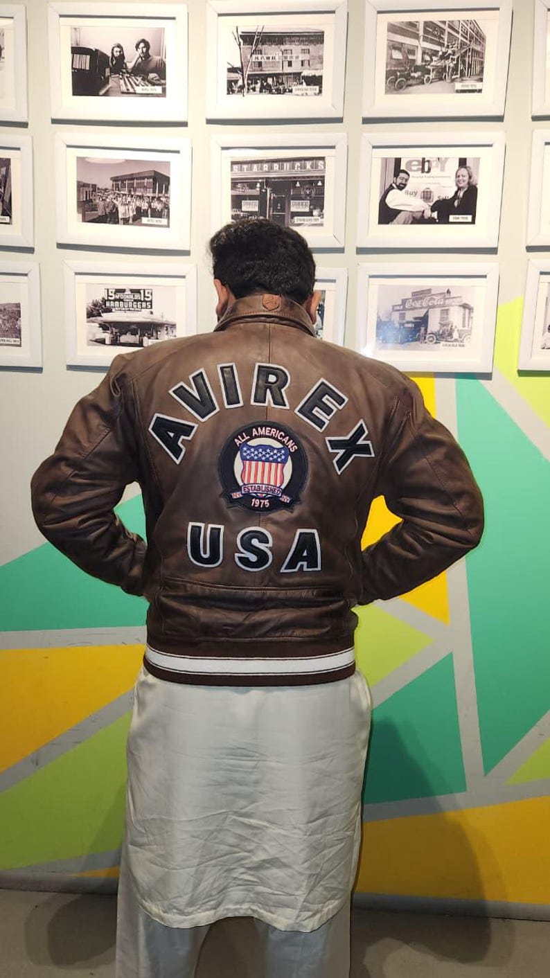 May include: A brown leather jacket with the word "AVIREX" in white lettering across the back, along with a patch that says "All Americans" and "USA". The jacket has a white and brown striped band at the waist.