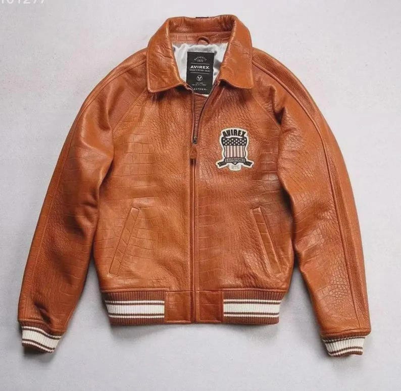 Pu&ograve; includere: Giacca in pelle marrone con motivo a coccodrillo. La giacca ha una chiusura con zip, un colletto e una toppa sul petto con la scritta "AVIREX LONDON". I polsini e la cintura hanno dettagli a righe bianche e marroni.