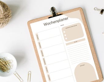 Puede incluir: Un planificador semanal en un portapapeles con el texto alemán "Wochenplaner". El planificador tiene secciones para cada día de la semana, una lista de tareas y espacio para notas. Clips dorados y un pequeño cuenco de clips están esparcidos.