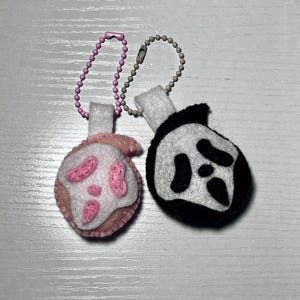 May include: Two handmade felt keychains. One is pink and white with pink accents, the other is black and white. Both have a small white tab and a ball chain with a clasp. The keychains are on a white surface.