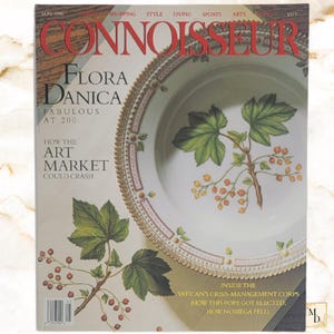 Op de afbeelding: Een uitgave van het tijdschrift Connoisseur uit mei 1990 met een wit porseleinen bord met een gouden rand en een botanische illustratie van groene bladeren en rode bessen. De tekst op de cover bevat Flora Danica en Art Market.