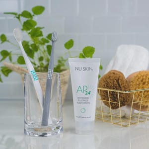 May include: A clear glass holds two toothbrushes, one white and one gray, next to a tube of AP24 Whitening Fluoride Toothpaste. A gold wire basket contains two brown bath sponges. Green foliage is in the background.