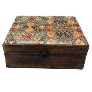 Può includere: Una scatola rettangolare in legno con finitura marrone scuro e coperchio incernierato. La superficie superiore presenta un motivo ripetitivo di forme intrecciate nei toni del rosso, verde acqua, oro e nero. Una piccola chiusura in metallo nero è centrata.