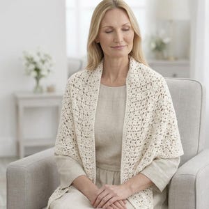 Puede incluir: Chal de ganchillo color crema con un diseño calado. El chal está drapeado sobre los hombros. La persona está sentada en un sillón gris, vistiendo un vestido de color claro. El fondo incluye una mesa con flores y una lámpara.