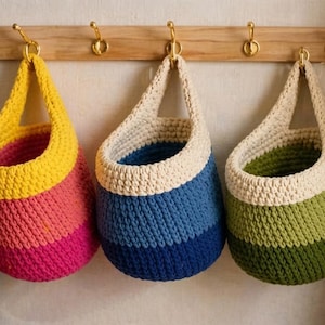 May include: Three hanging crochet baskets in shades of yellow, pink, blue, and green. Each basket is suspended from a wooden rack with brass hooks. The baskets have a rounded shape and are designed for storage or decoration.