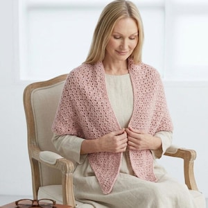 Może przedstawiać: Jasnoróżowy szydełkowy szal z teksturowanym wzorem. Szal jest narzucony na ramiona i ma trójkątny kształt z przodu. Kobieta siedzi na beżowym krześle.