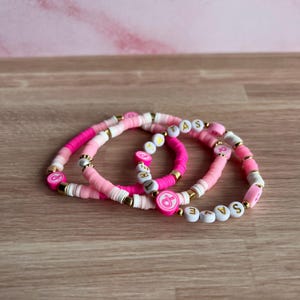 May include: Three pink and white beaded bracelets with gold accents. One bracelet has the word "SAVE" spelled out in white letter beads. Another bracelet has the word "TAS" spelled out in white letter beads. Each bracelet has a pink ribbon charm.