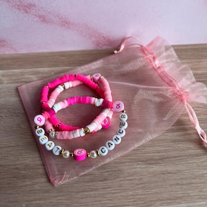 May include: Three pink and white beaded bracelets with the words "BEAT CANCER" spelled out in white letter beads. The bracelets are on a pink organza bag, with a pink background.