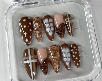 May include: A set of ten almond-shaped press-on nails in a clear plastic case. The nails feature various designs, including white polka dots on brown, marbled patterns, coffee bean accents, and white crosshatch designs. Some nails are a nude pink color.