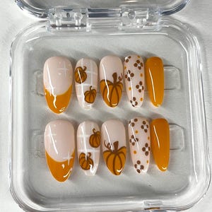 May include: A set of ten artificial nails in a clear plastic case. The nails feature a light pink base with orange tips and pumpkin designs. Some nails have a pumpkin graphic, while others have a polka dot pattern.