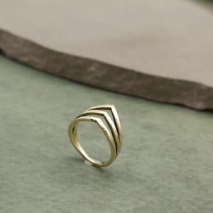 May include: A gold-colored ring with a layered chevron design. The ring features three stacked V-shaped bands, creating a geometric and modern aesthetic. The ring is set against a muted green and gray background.