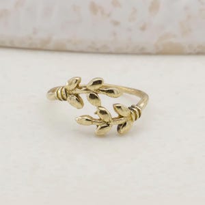 May include: A gold-coloured ring with a leaf design. The ring features two branches of leaves that wrap around the finger. The ring is made of a shiny, metallic material and is set against a neutral background.