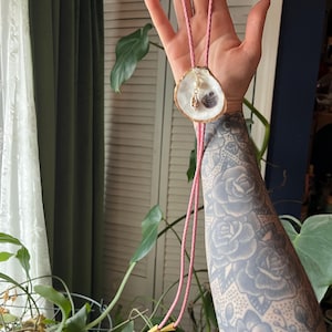 May include: A bolo tie featuring a pink braided cord with gold-toned metal tips. The central focal point is a white oyster shell with a gold-painted edge, accented by a small dangling charm consisting of a white pearl and a tiny spiral seashell.