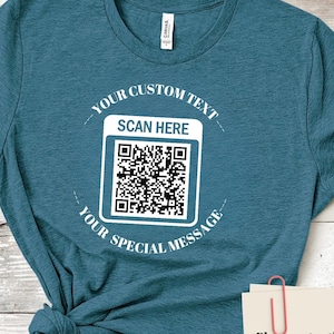 May include: Teal t-shirt with a QR code graphic. The shirt has the text "YOUR CUSTOM TEXT" above the QR code and "YOUR SPECIAL MESSAGE" below it. The text "SCAN HERE" is above the QR code. A note attached with a paper clip provides instructions for the QR code.