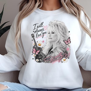 Puede incluir: Una sudadera blanca de manga larga con un gráfico en blanco y negro de una mujer de pelo rubio, una guitarra acústica negra, margaritas y mariposas rosas. El texto dice I will always love you y Dolly, con corazones y estrellas rosas.
