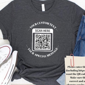May include: Dark grey heather t-shirt with a white QR code graphic. The text around the QR code reads "YOUR CUSTOM TEXT" and "YOUR SPECIAL MESSAGE". The shirt also has the text "SCAN HERE".