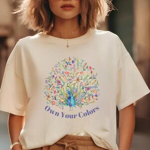 May include: Cream-colored t-shirt featuring a colorful peacock design with the text "Own Your Colors" in blue. The peacock is surrounded by floral and botanical elements in shades of blue, green, and purple.