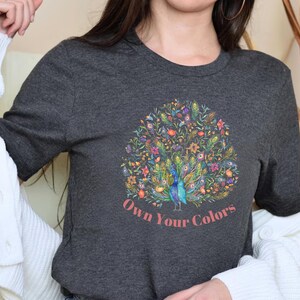 May include: Dark gray t-shirt featuring a colorful peacock design with floral accents. The peacock is depicted in shades of blue and green, surrounded by flowers in various colors. The text "Own Your Colors" is printed in a pinkish-red font below the peacock.