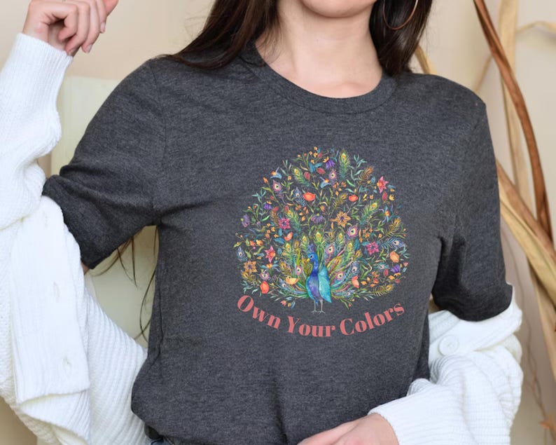 May include: Dark gray t-shirt featuring a colorful peacock design with floral accents. The peacock is depicted in shades of blue and green, surrounded by flowers in various colors. The text "Own Your Colors" is printed in a pinkish-red font below the peacock.