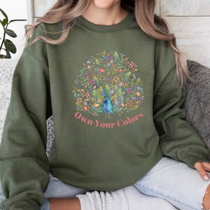 May include: Olive green sweatshirt featuring a colorful peacock design with floral accents. The peacock is in the center, with the words "Own Your Colors" in a script font below the design. The sweatshirt has long sleeves.