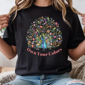 May include: Black t-shirt featuring a colorful peacock design with the text "Own Your Colors". The peacock is depicted with vibrant blue, green, yellow, and pink feathers, surrounded by floral elements. The text is in a pink script font.