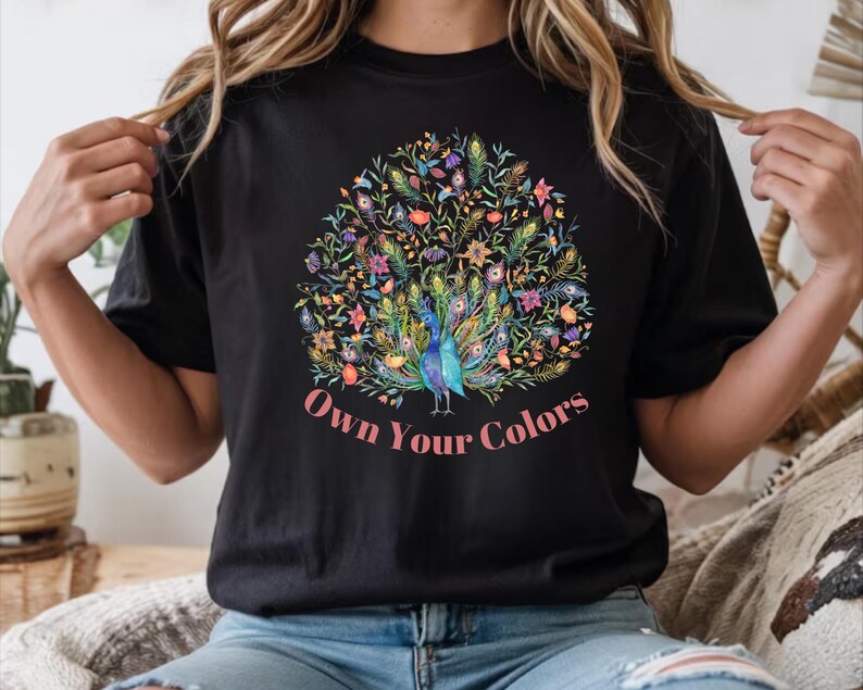 May include: Black t-shirt featuring a colorful peacock design with the text "Own Your Colors". The peacock is depicted with vibrant blue, green, yellow, and pink feathers, surrounded by floral elements. The text is in a pink script font.