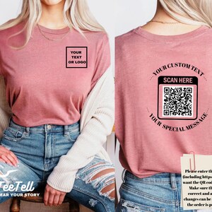 May include: A dusty rose t-shirt with a square box that says "YOUR TEXT OR LOGO" on the front. The back of the shirt has a QR code with the text "YOUR CUSTOM TEXT SCAN HERE YOUR SPECIAL MESSAGE". The shirt is paired with blue jeans.