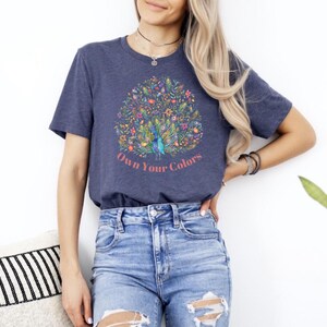 May include: A navy blue t-shirt with a colorful peacock design and the text "Own Your Colors." The peacock design features a variety of flowers and plants. The shirt is paired with blue denim jeans.