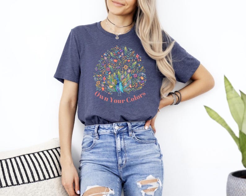 May include: A navy blue t-shirt with a colorful peacock design and the text "Own Your Colors." The peacock design features a variety of flowers and plants. The shirt is paired with blue denim jeans.