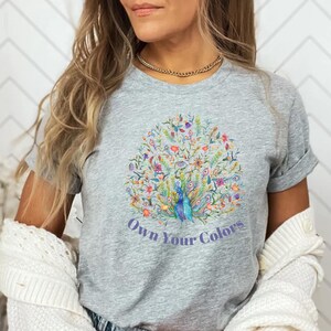 May include: A gray t-shirt featuring a colorful peacock design with the text "Own Your Colors". The peacock design includes blue, green, red, and yellow details. The shirt has short sleeves and a crew neck.