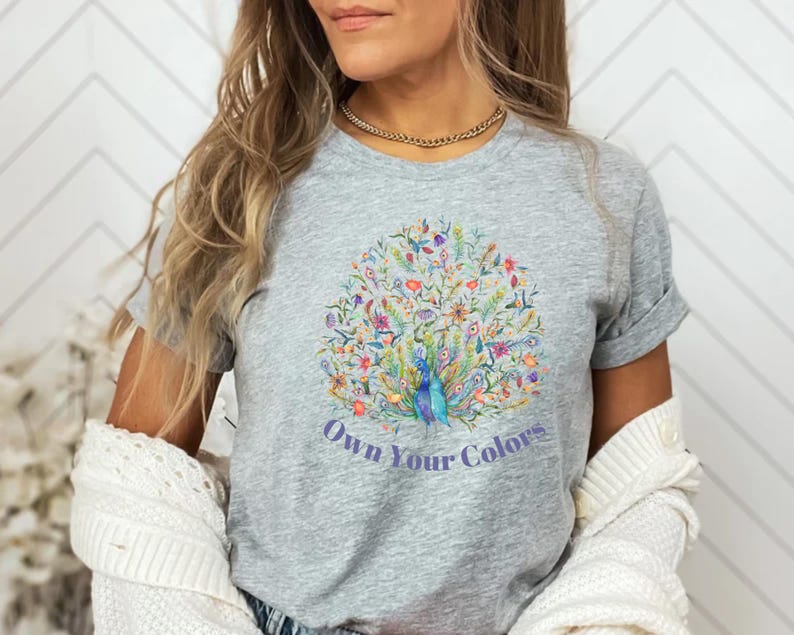 May include: A gray t-shirt featuring a colorful peacock design with the text "Own Your Colors". The peacock design includes blue, green, red, and yellow details. The shirt has short sleeves and a crew neck.