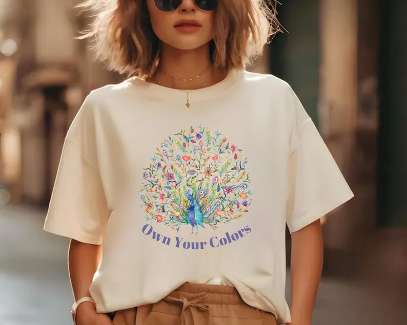 May include: Cream-colored t-shirt featuring a colorful peacock design with the text "Own Your Colors" in blue. The peacock is surrounded by floral and botanical elements in shades of blue, green, and purple.