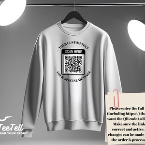 May include: White sweatshirt with a crew neck, hanging on a black hanger. The front features a QR code with the text "YOUR CUSTOM TEXT", "SCAN HERE", and "YOUR SPECIAL MESSAGE". A paper with instructions is clipped to the sweatshirt.