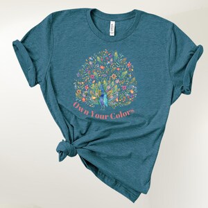 May include: Teal-colored t-shirt featuring a colorful peacock design with floral accents. The peacock is surrounded by flowers and foliage. The text "Own Your Colors" is printed below the peacock in a pink font. The t-shirt is knotted at the bottom.