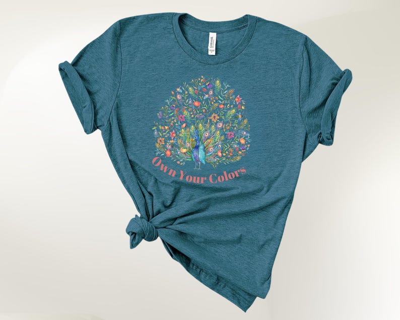 May include: Teal-colored t-shirt featuring a colorful peacock design with floral accents. The peacock is surrounded by flowers and foliage. The text "Own Your Colors" is printed below the peacock in a pink font. The t-shirt is knotted at the bottom.