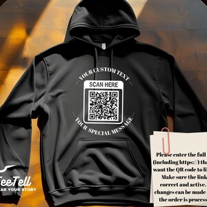 May include: Black hoodie with a front pocket and a hood. The front features a white graphic with a QR code and the text "YOUR CUSTOM TEXT SCAN HERE YOUR SPECIAL MESSAGE". A note with instructions is clipped to the hoodie.