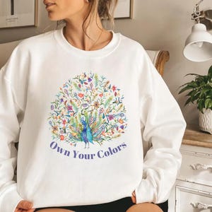 May include: White crew neck sweatshirt with a colorful peacock design. The peacock is surrounded by flowers and foliage in shades of blue, green, yellow, and purple. The text "Own Your Colors" is printed below the peacock.