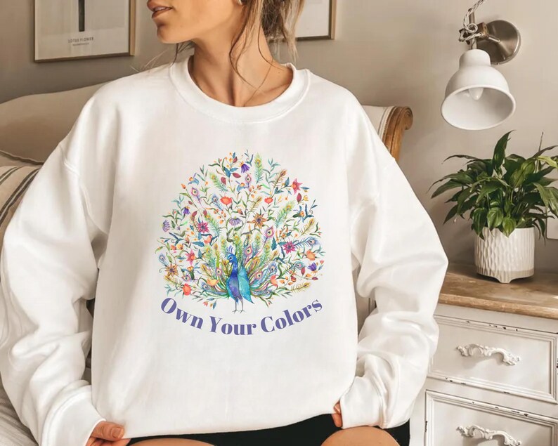 May include: White crew neck sweatshirt with a colorful peacock design. The peacock is surrounded by flowers and foliage in shades of blue, green, yellow, and purple. The text "Own Your Colors" is printed below the peacock.