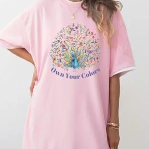 May include: A light pink t-shirt dress with a white trim on the sleeves. The front features a colorful peacock design with the text "Own Your Colors" in a blue font.