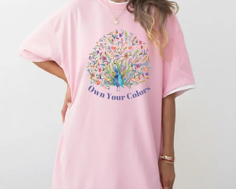 May include: A light pink t-shirt dress with a white trim on the sleeves. The front features a colorful peacock design with the text "Own Your Colors" in a blue font.