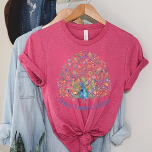 May include: A magenta t-shirt with a watercolor peacock design. The peacock has blue, green, and yellow feathers, surrounded by colorful floral elements. The text "Own Your Colors" is below the peacock.