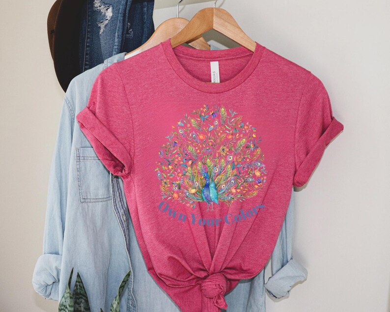 May include: A magenta t-shirt with a watercolor peacock design. The peacock has blue, green, and yellow feathers, surrounded by colorful floral elements. The text "Own Your Colors" is below the peacock.