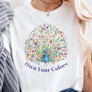 May include: White t-shirt featuring a colorful peacock design with the text "Own Your Colors". The peacock is surrounded by flowers and leaves in shades of blue, green, yellow, and purple. The shirt is a casual wear item.